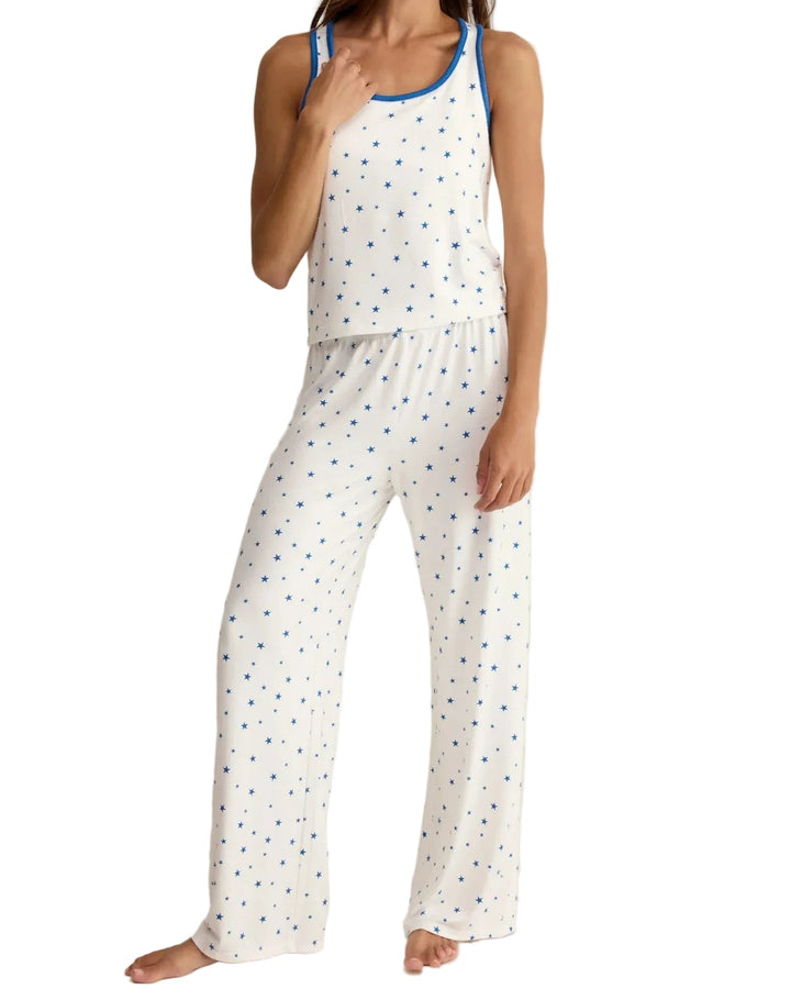 A woman modeling a white sleeveless pajama set with a small blue polka dot pattern and blue trim on the tank top, photographed against a plain light beige studio background.