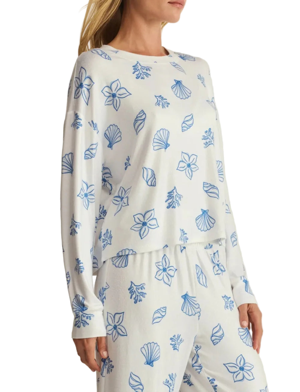 A woman modeling a white long-sleeve pajama top and matching pants set with a blue tropical floral and shell pattern, set against a plain beige background.