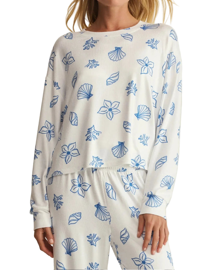 A woman wearing a matching long-sleeve pajama set with a white base and blue seashell and floral line-art print, photographed against a plain light beige studio background.