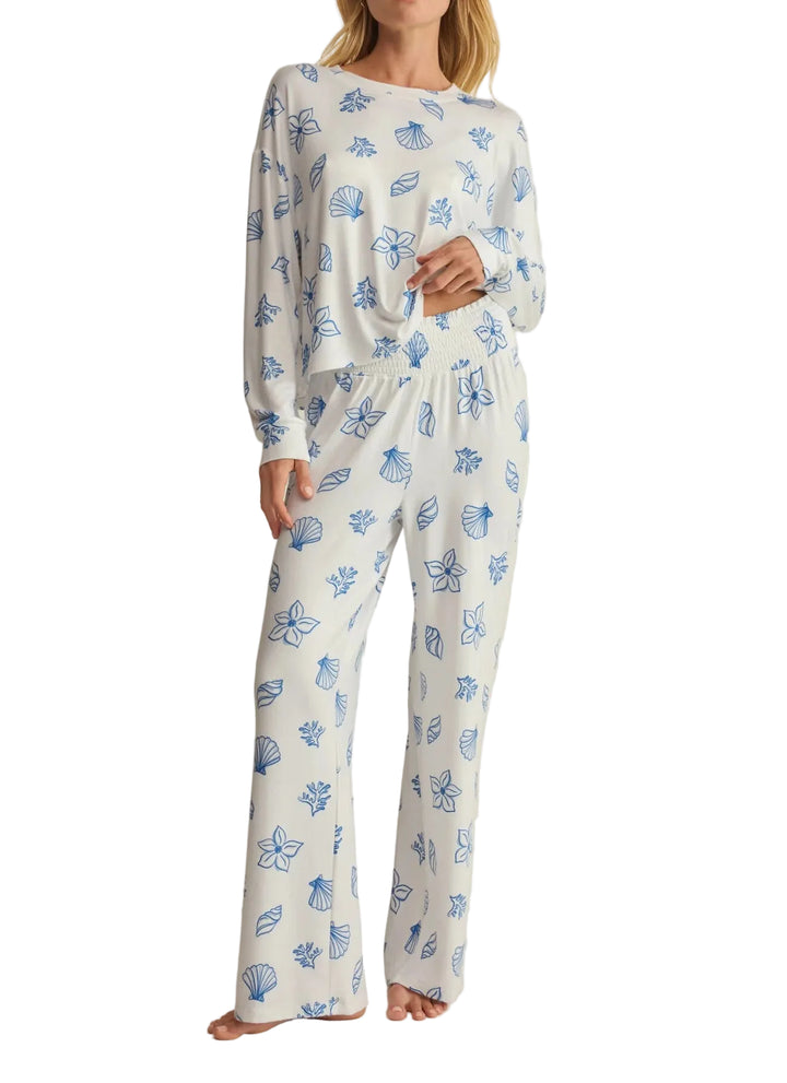 A woman modeling a two-piece long-sleeve pajama set with a white base and blue floral and shell-like print, photographed against a plain light beige studio background.