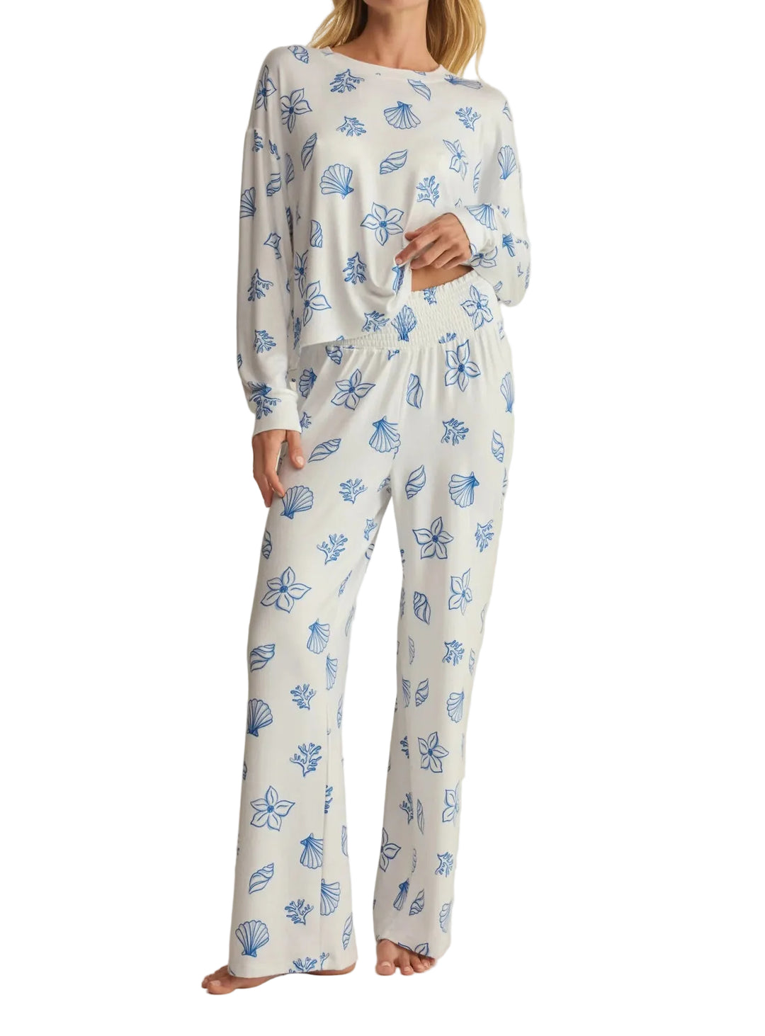 A woman modeling a two-piece long-sleeve pajama set with a white base and blue floral and shell-like print, photographed against a plain light beige studio background.