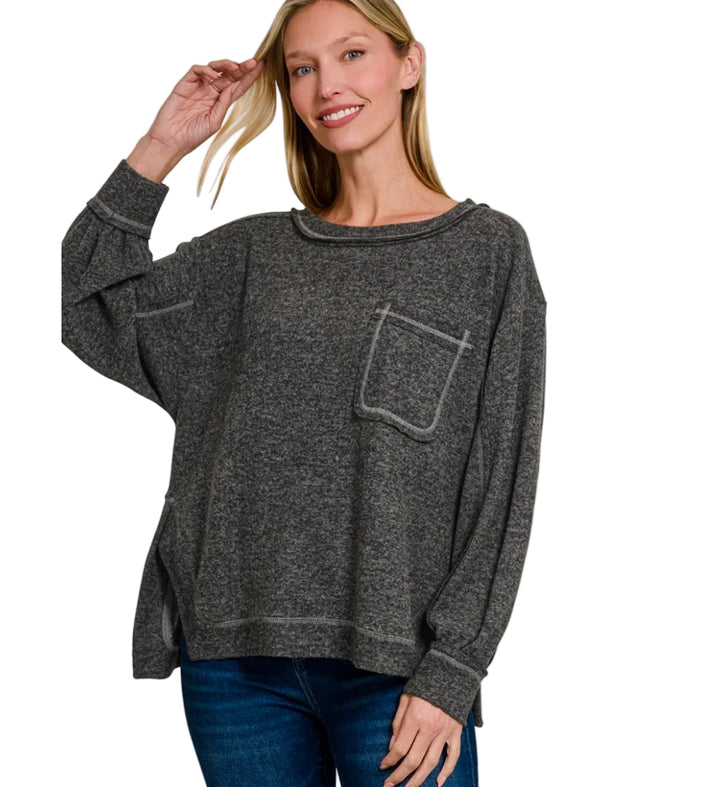 A woman wearing a dark heather gray oversized crewneck sweater with a chest pocket, paired with blue jeans, against a plain white background.  'BRUSHED SOFT' and 'BLACK'.