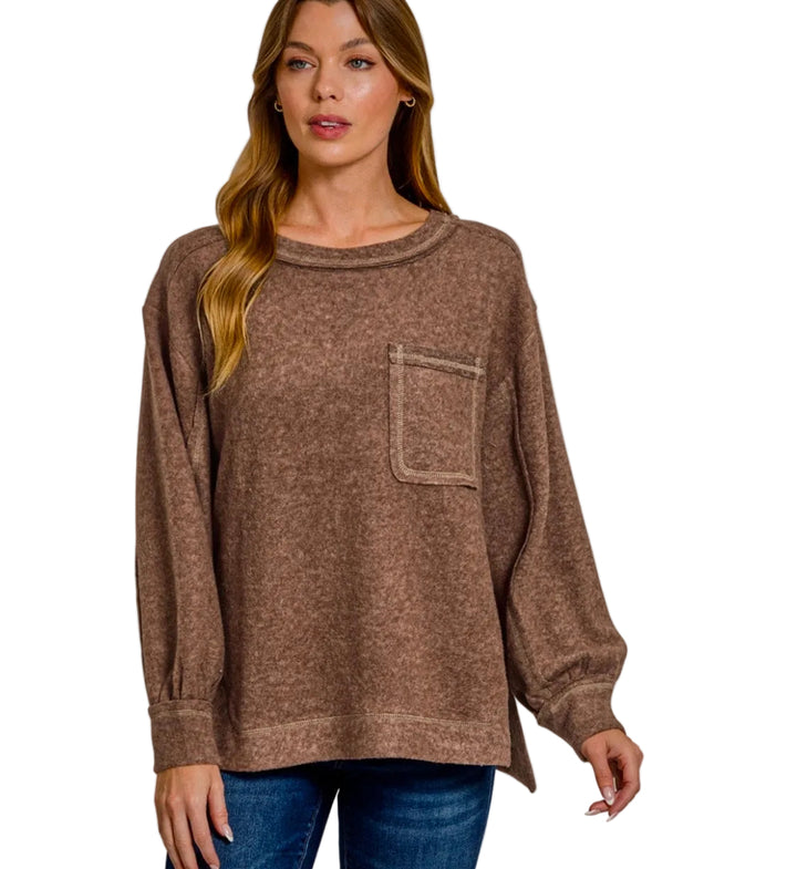 A woman modeling a brown long-sleeve knit sweater with a chest pocket, paired with blue jeans, against a light gray background.