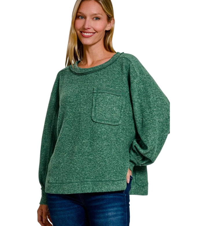 A woman wearing a heathered dark green oversized crewneck sweatshirt with a chest pocket, paired with blue jeans, against a plain light gray background.