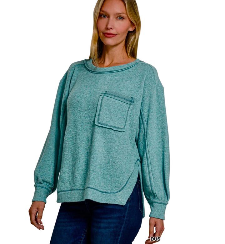A woman modeling a dusty teal heathered sweatshirt with a chest pocket, paired with dark blue jeans, against a light gray background.