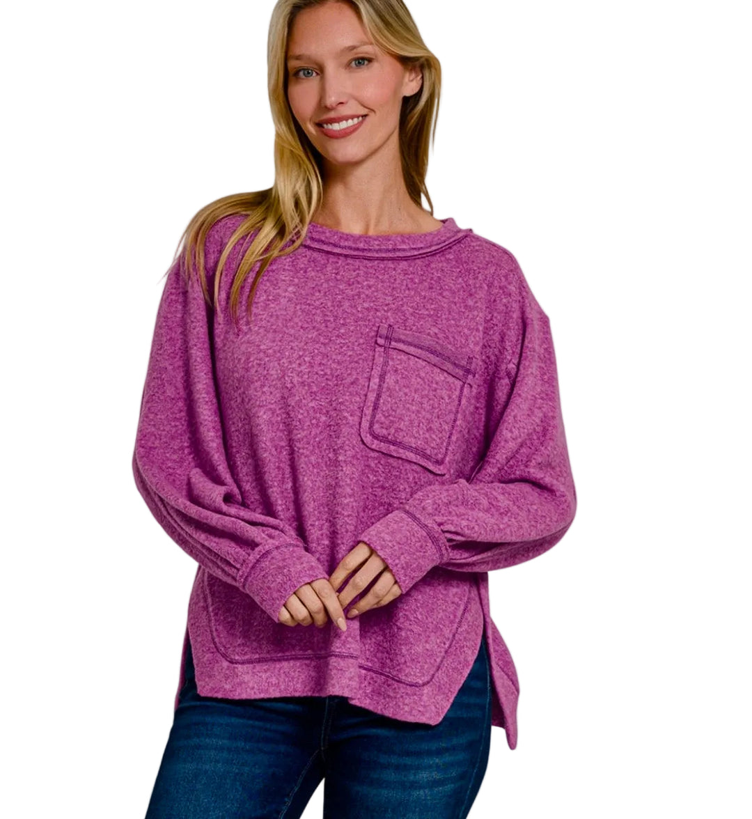 A woman wearing a heathered plum purple long-sleeve knit top with a chest pocket, paired with dark blue jeans, against a light gray background. Text overlays read 'BRUSHED WEATER SOFT' and 'LT PLUM'.