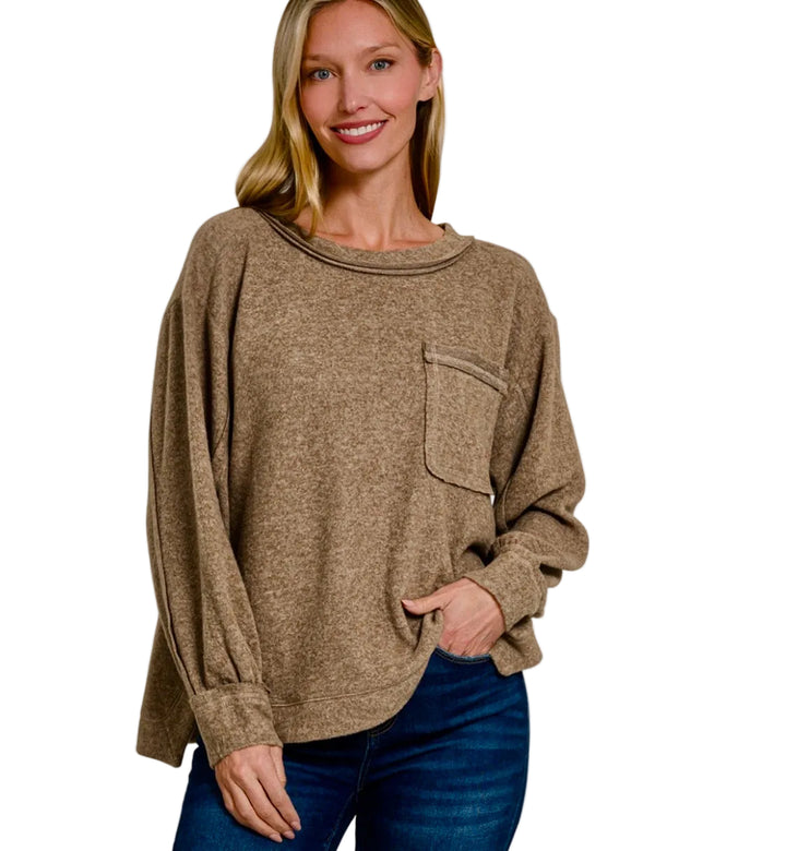 A woman wearing a loose, long-sleeve knit top in a warm taupe-brown color with a chest pocket, paired with blue jeans, against a light gray background. Text overlays read 'BRUSHED MIDWEATER SOFT' and 'MOCHA'.