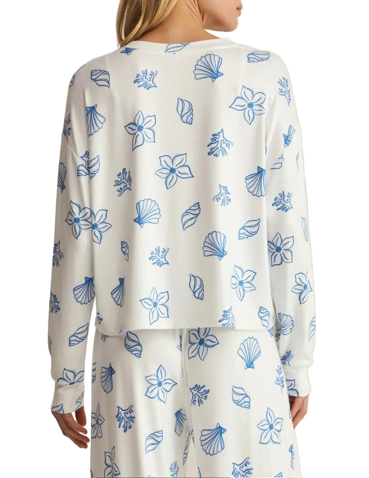 A woman shown from the back wearing a white long-sleeve top and matching pants set, both printed with a blue floral and shell-like line pattern, against a plain light beige studio background.