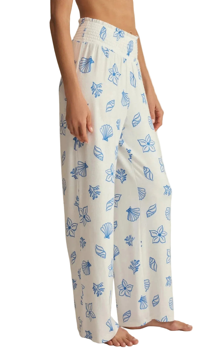 White wide-leg lounge pants with a blue seashell and floral pattern, shown on a model against a warm beige background.